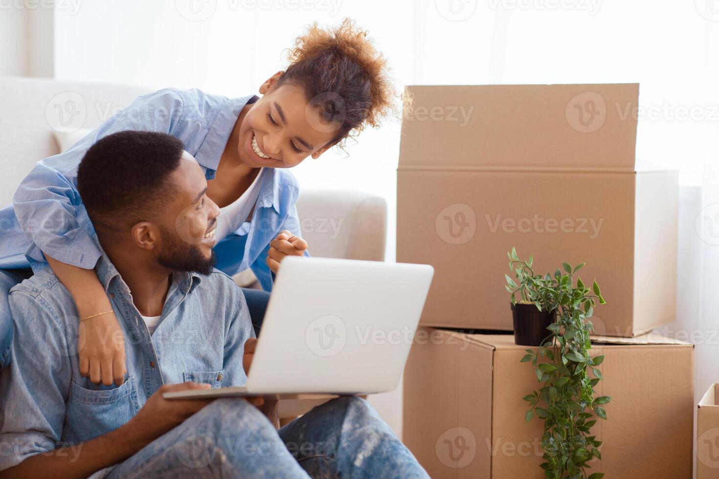 Excited Afro Spouses Using Laptop Computer Sitting On Sofa Among Unpacked Moving Boxes In New House. Free Space, Selective Focus photo