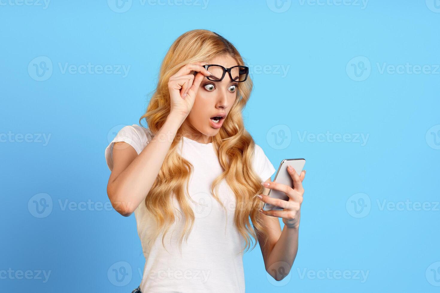This image shows a surprised young woman with blonde hair wearing glasses looking at a smartphone with a shocked expression on her face. She is standing in front of a solid blue background. photo