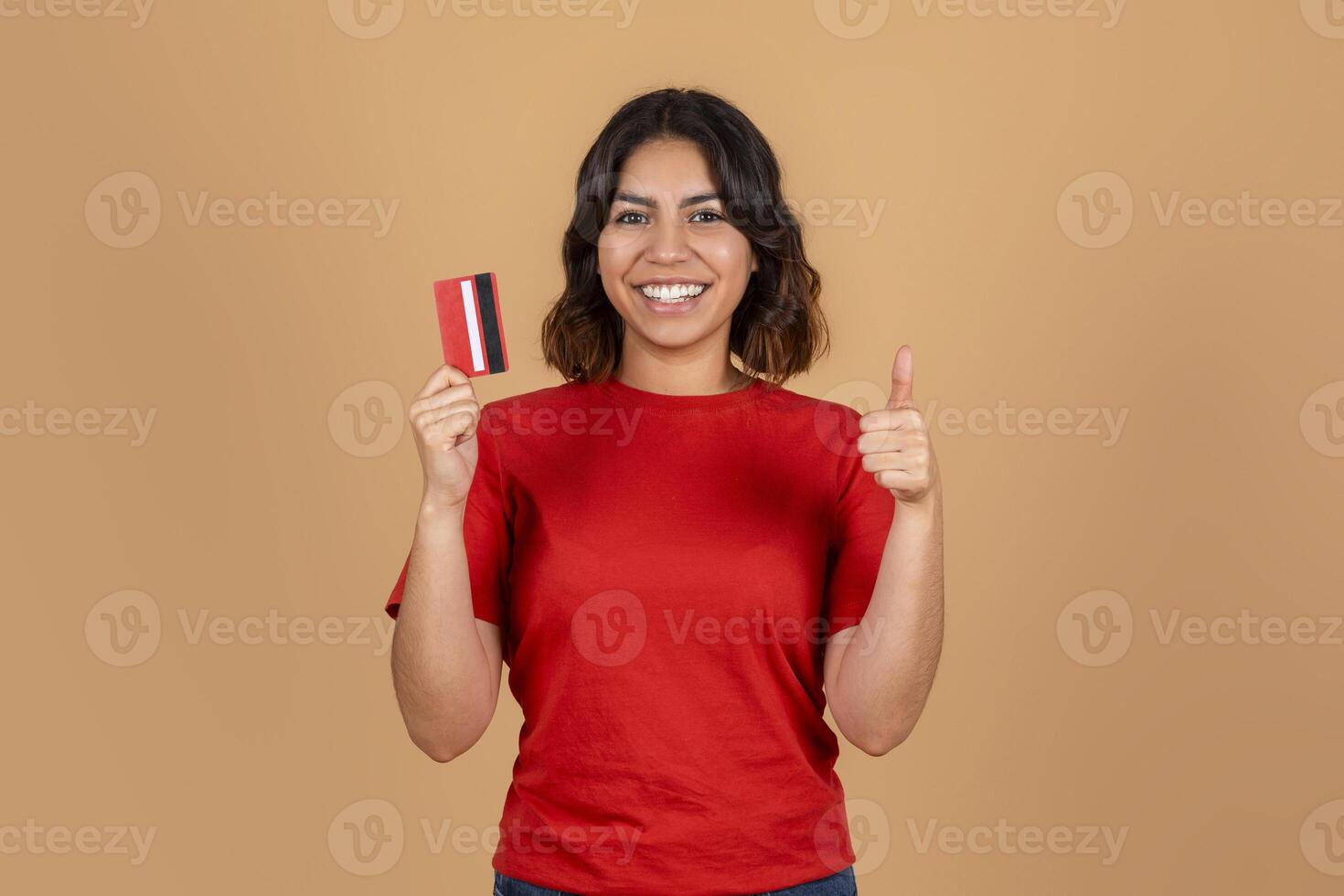 A middle eastern woman in a red t-shirt stands in front of a light brown background. She is holding a red credit card in her left hand and giving a thumbs up with her right hand photo