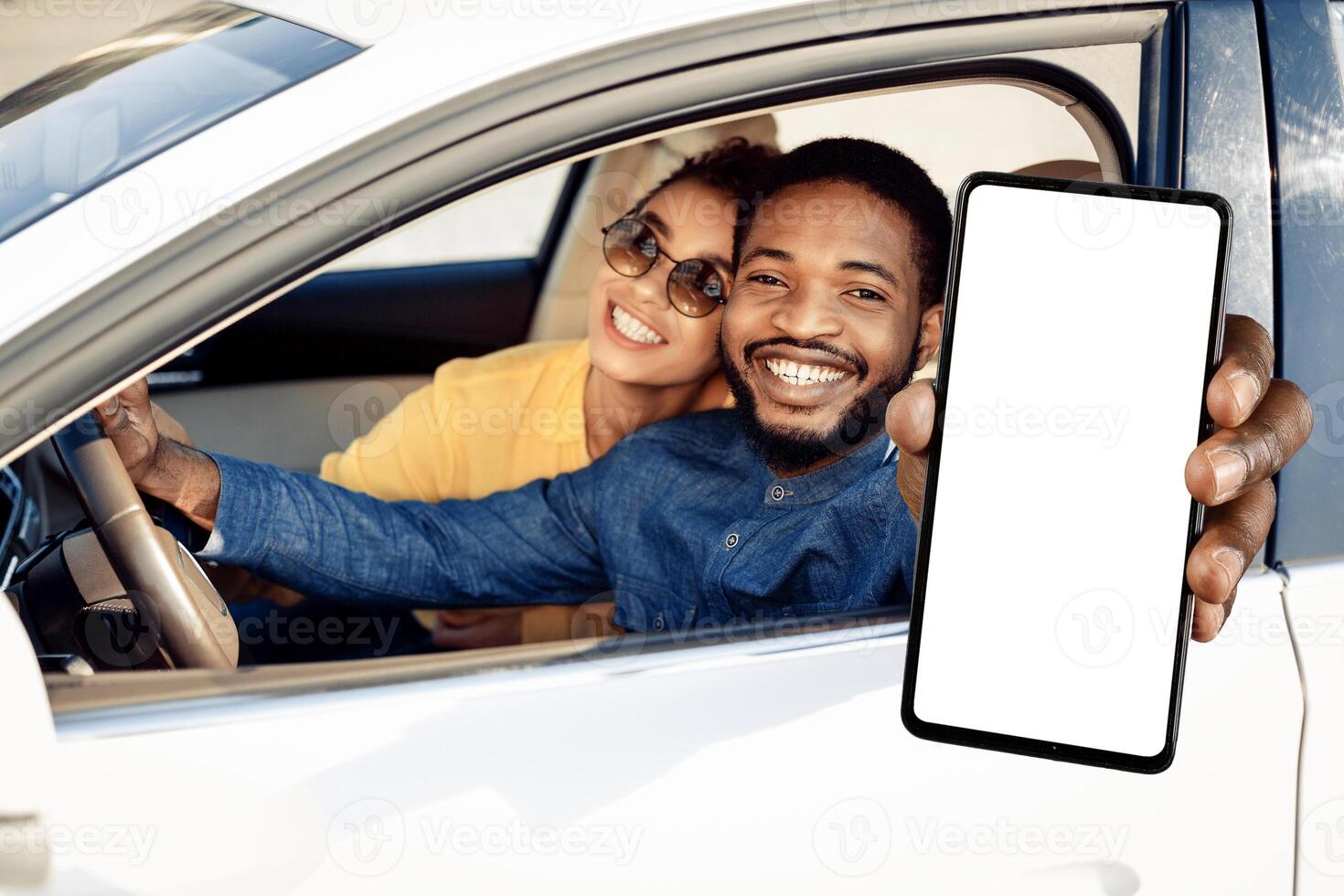 A smiling young black man sits in the drivers seat of a car, holding a smartphone with a blank white screen out the window. A smiling young woman sits beside him. photo