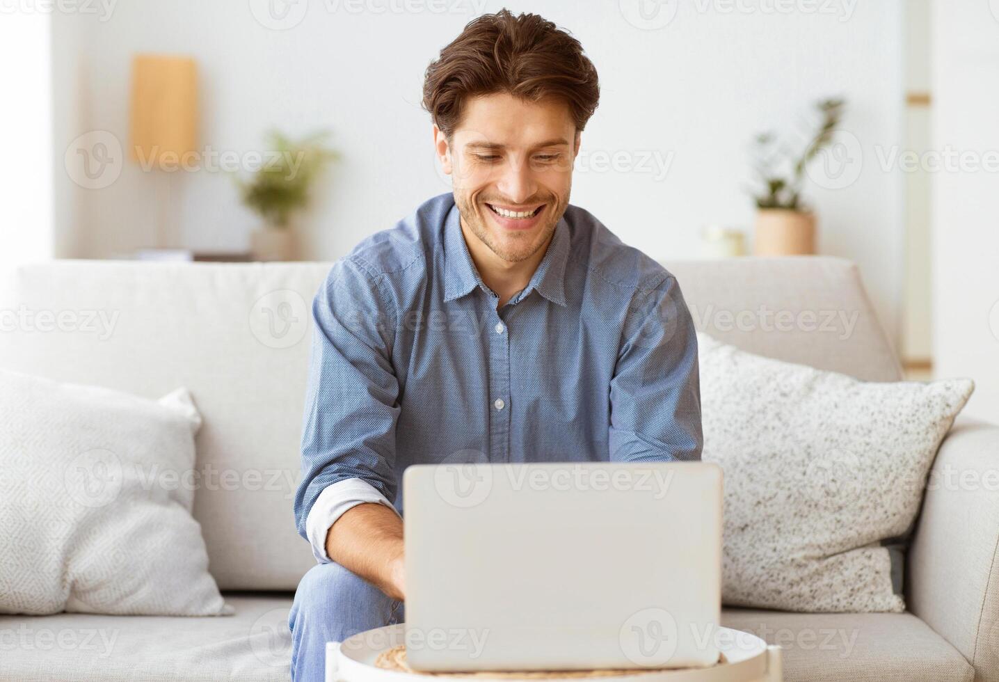 A man sits on a couch and smiles while using a laptop. photo