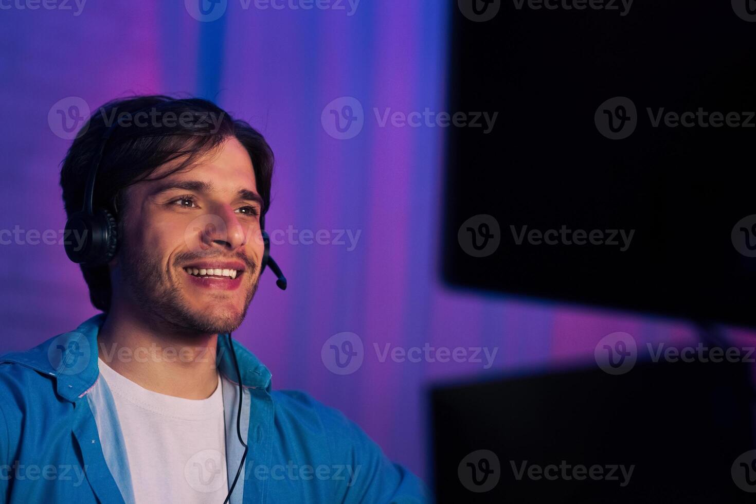 A young man smiles while looking off to the side. He is wearing a blue shirt and a headset. He is sitting in front of a computer with a purple and blue backdrop. photo