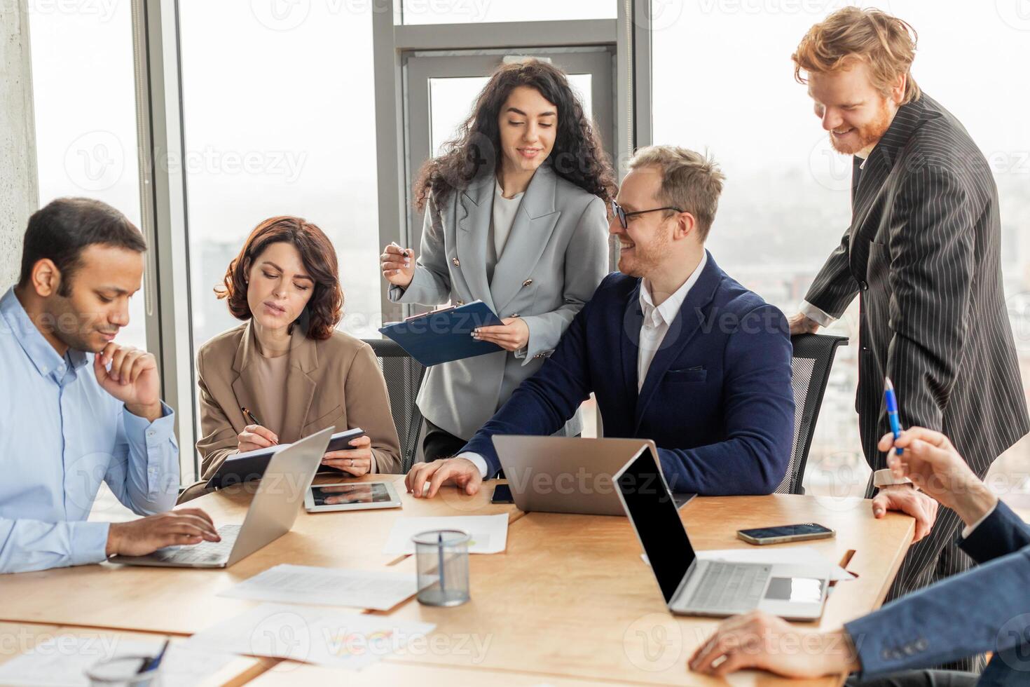 A group of business professionals, including men and women, work together at a large wooden table in a modern office. They are looking at laptops, taking notes, and discussing ideas. photo