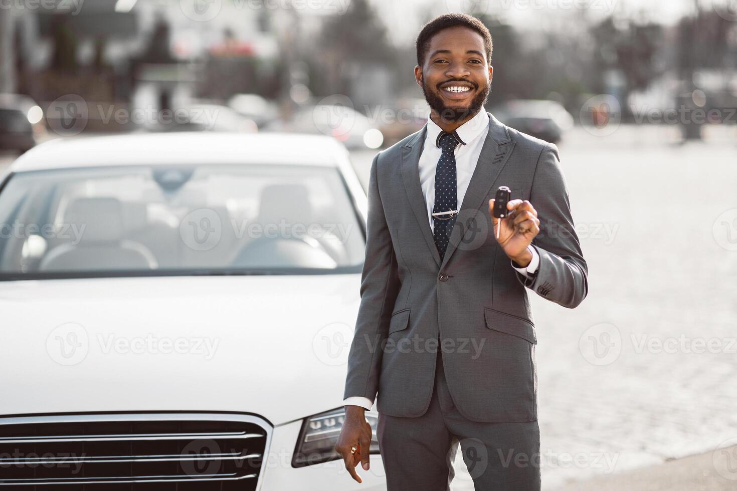 This image shows African American man in a grey suit, smiling, holding a car key. He is standing in front of a new white car. He looks proud and happy. photo