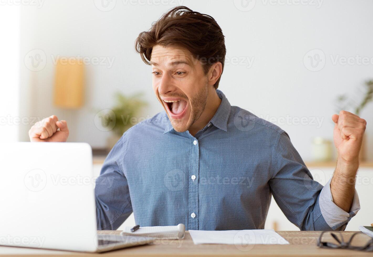 A man sits at a desk, looking at his laptop with a surprised, excited expression. He is celebrating his success with raised fists. photo