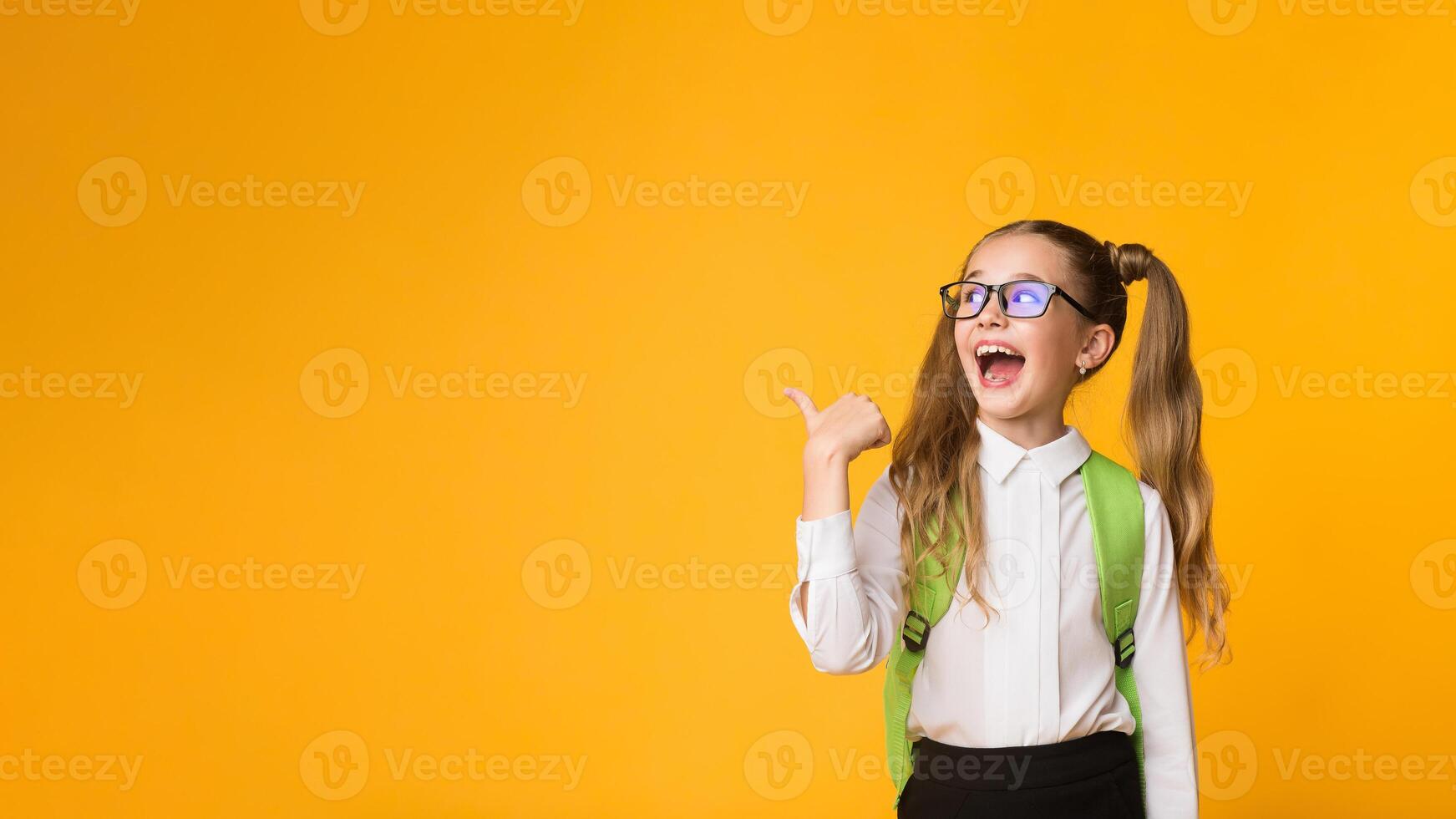Look There. Excited Elementary Student Girl Pointing Thumb At Free Space Over Yellow Background In Studio. Panorama photo