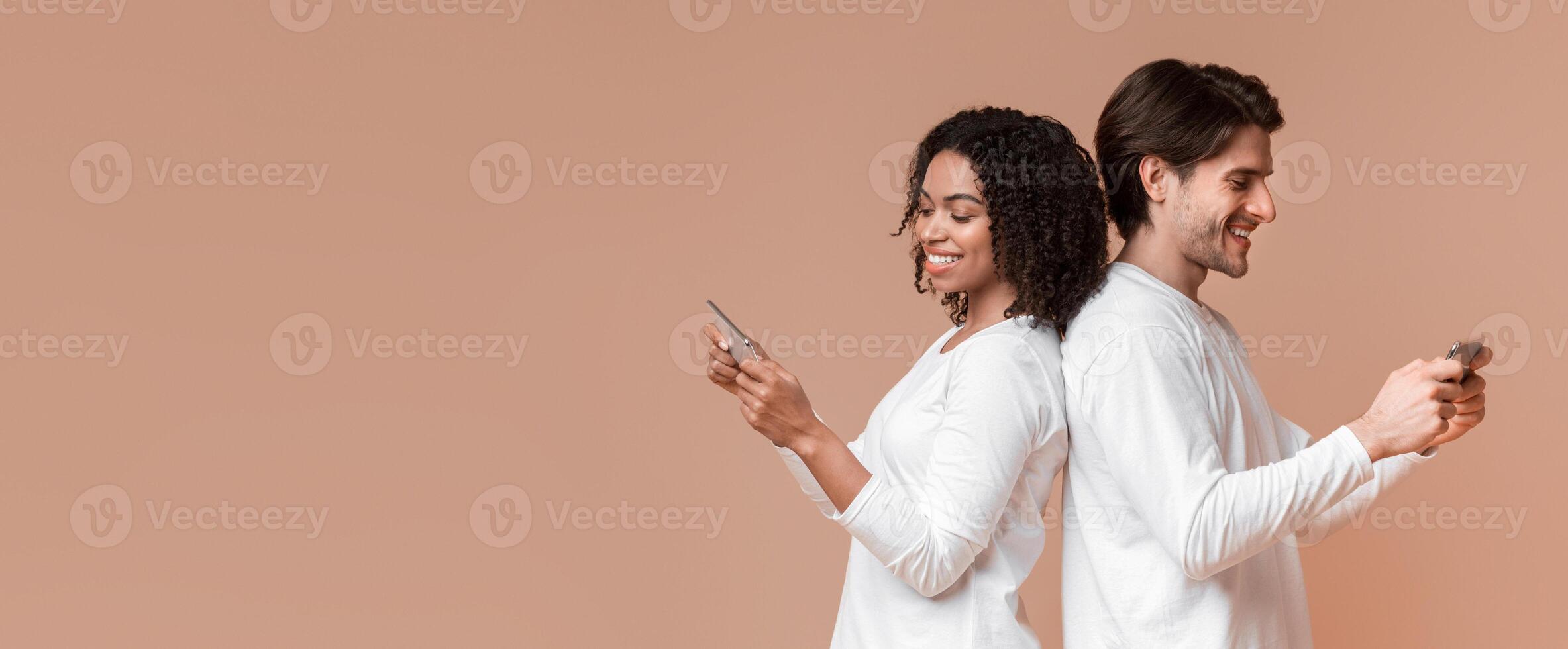 Gadget and social media addiction concept. Smiling multiracial couple standing back to back and using their smartphones, ignoring each other photo