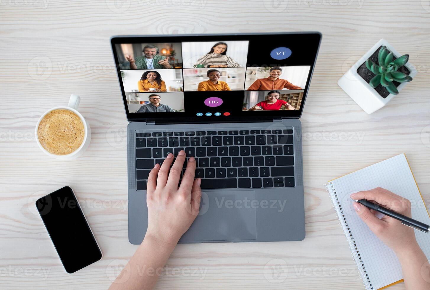 A woman is participating in a conference meeting on her laptop. She is typing on the keyboard with one hand and writing notes with the other. photo