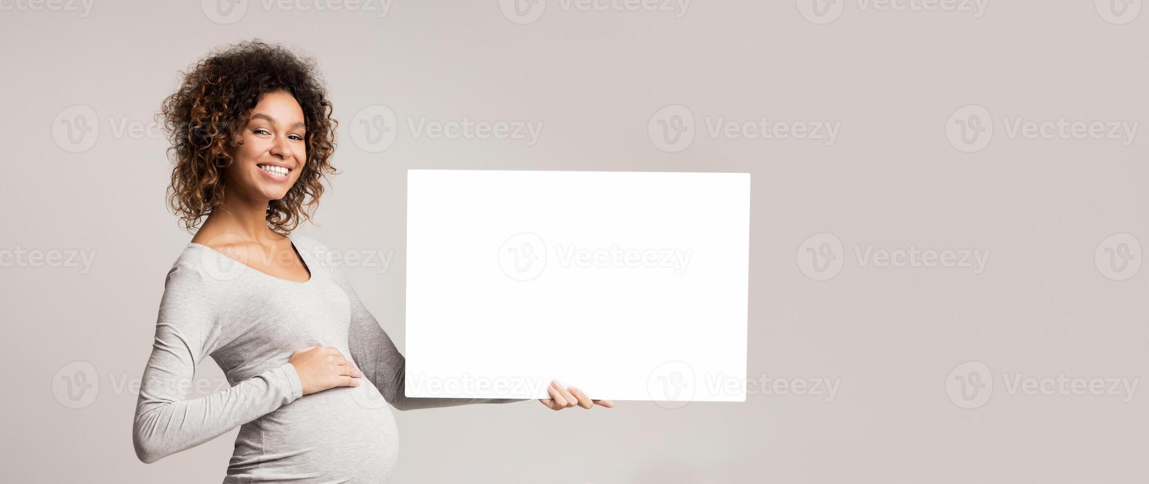 Black expectant woman holding blank advertising board over gray studio background, free space photo