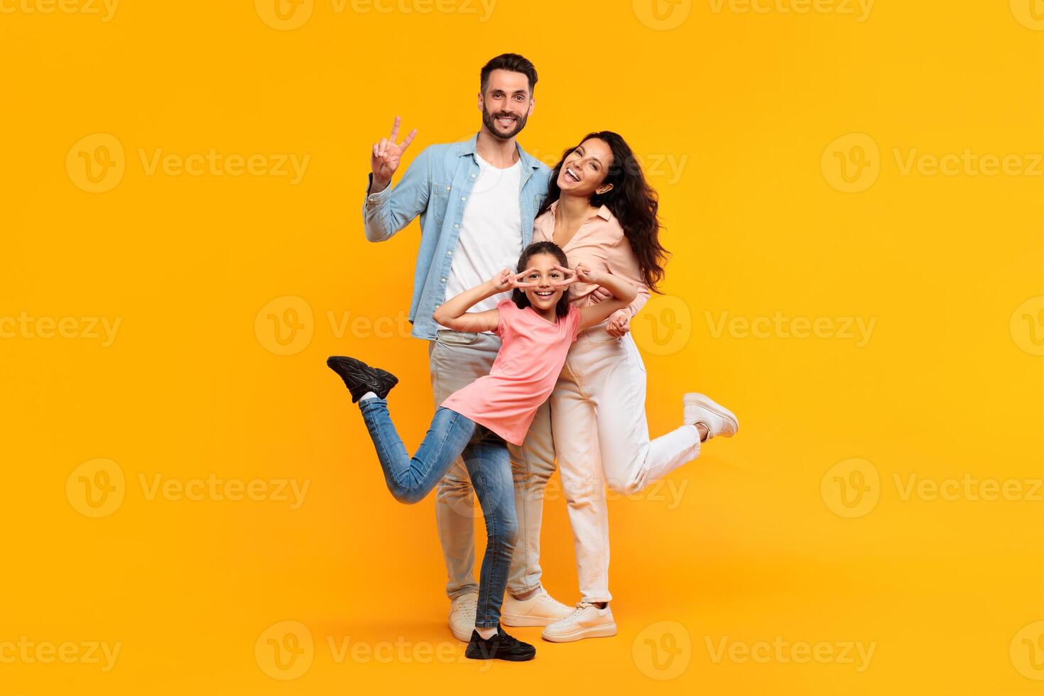 A happy family of three poses together in front of a yellow background. The father is on the left, making a peace sign with his hand, while the mother is on the right, with a bright smile. photo