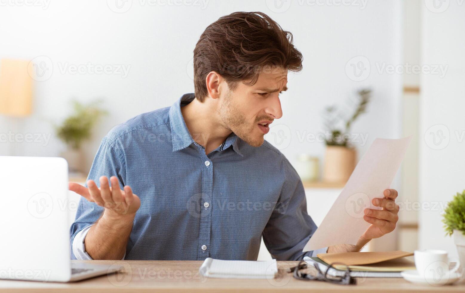 A man sits at a desk looking perplexed while reading a document. His expression suggests he is unhappy or surprised by the contents. photo