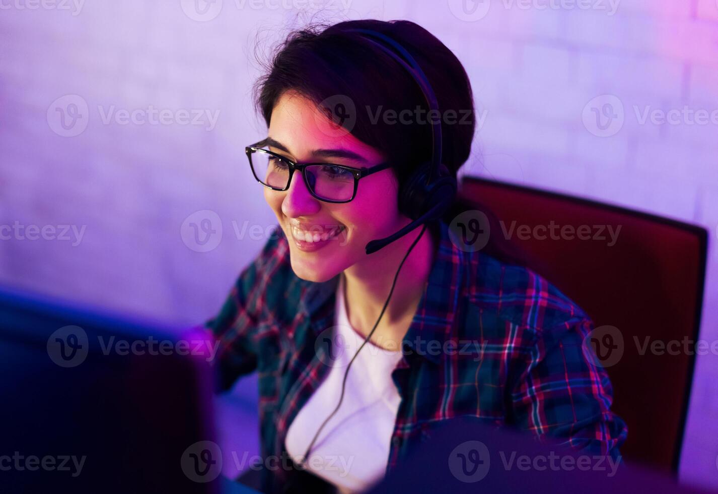 A young woman with dark hair is wearing glasses and a headset while sitting in front of a computer. She is smiling and looking at the screen. The room is lit in a purple and pink glow photo