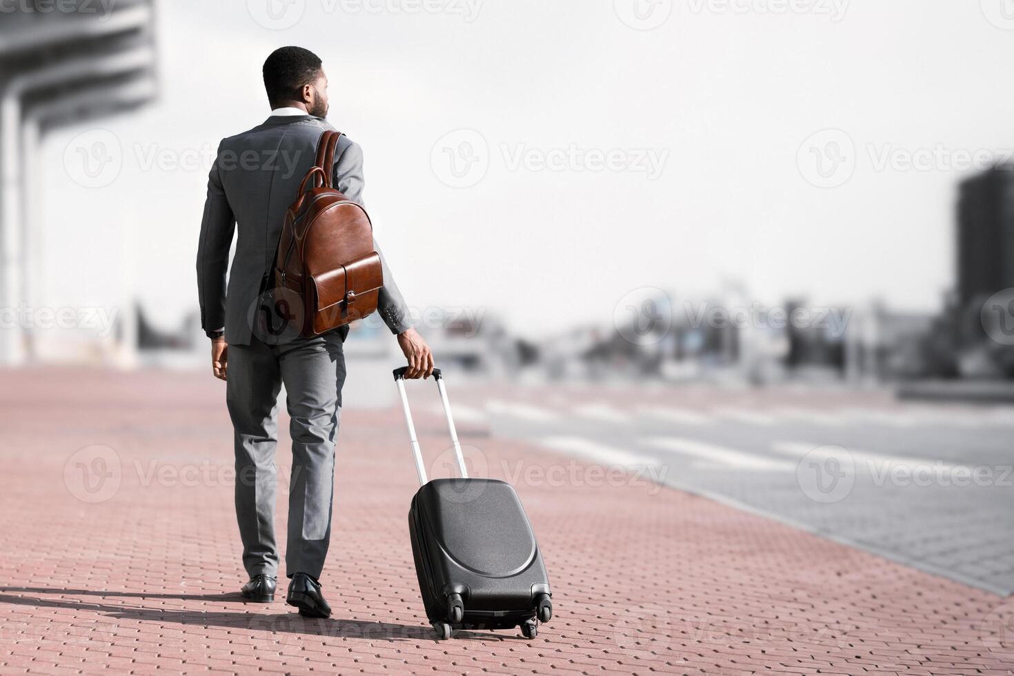 Back view of black businessman walking with rolling suitcase outside a modern building, ready for a business trip or travel. photo