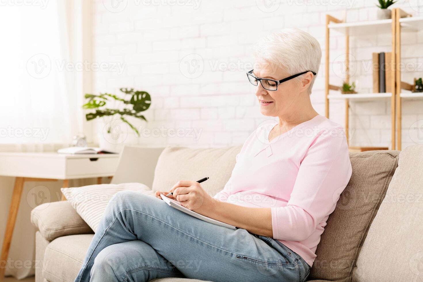Senior woman is seated on a couch, concentrating as she writes on a piece of paper placed on her lap. Her focus is intense, highlighting her engagement in the task at hand. photo