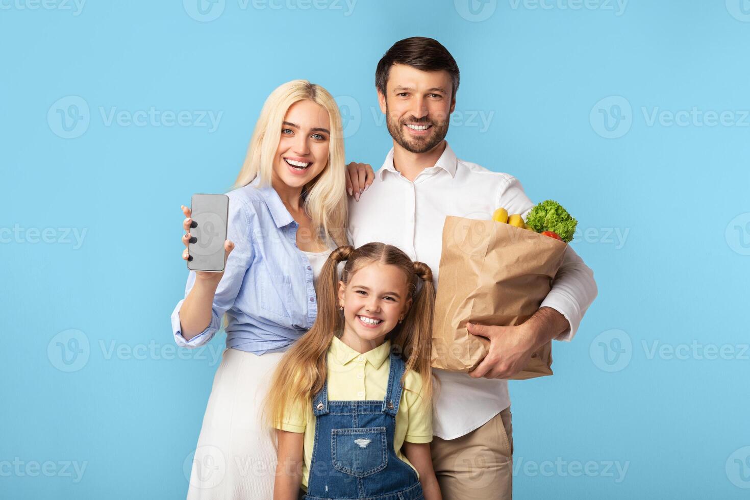 A happy family of three stands in front of a light blue background. The mother holds a smartphone in her right hand and smiles at the camera. The father holds a paper bag full of groceries photo