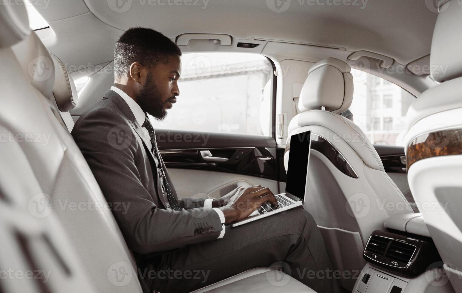 African American businessman sits in the backseat of a car, working on his laptop computer. He is wearing a suit and tie and appears to be focused on his work photo