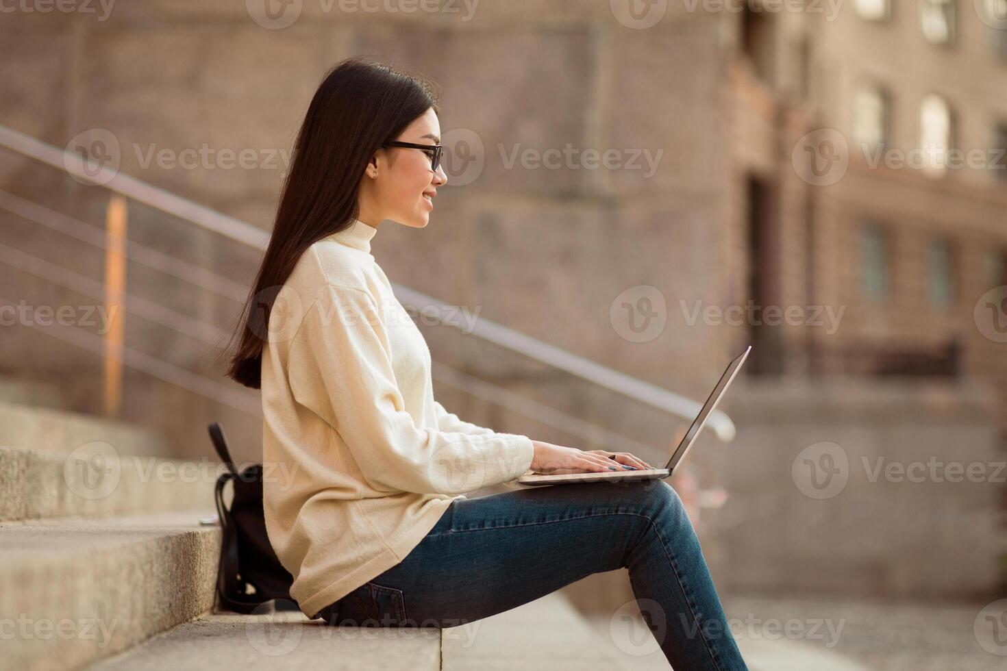 Asian woman student is sitting on a flight of steps, focused on her laptop screen. She appears engrossed in her work or studies, with buildings in the background. photo