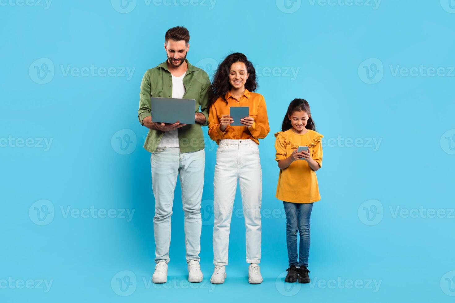 A man, woman, and young girl stand in a row against a light blue background. Family using various devices. Gadgets addiction photo