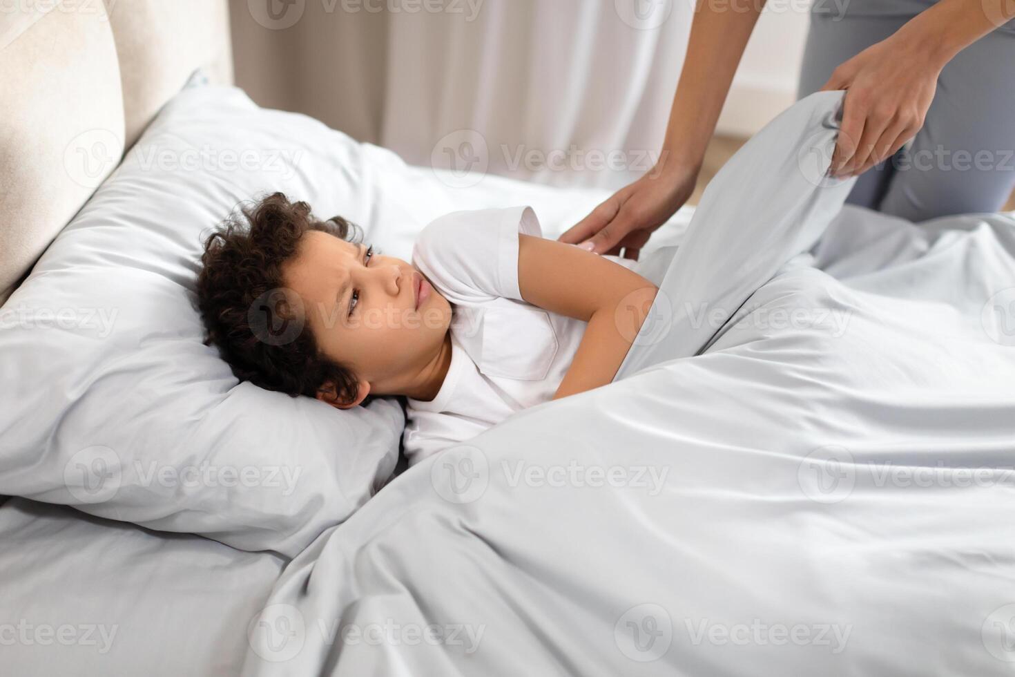 African American young child rests on a bed with a look of anticipation, watching as a parent lovingly adjusts the blanket over them photo