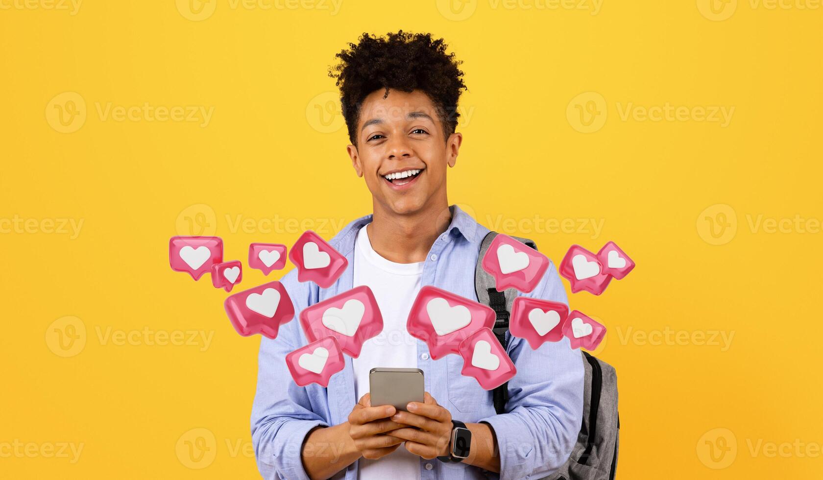Cheerful young black man student with backpack comfortably using his smartphone, texting girlfriend, smiling at the camera standing against vivid yellow backdrop photo