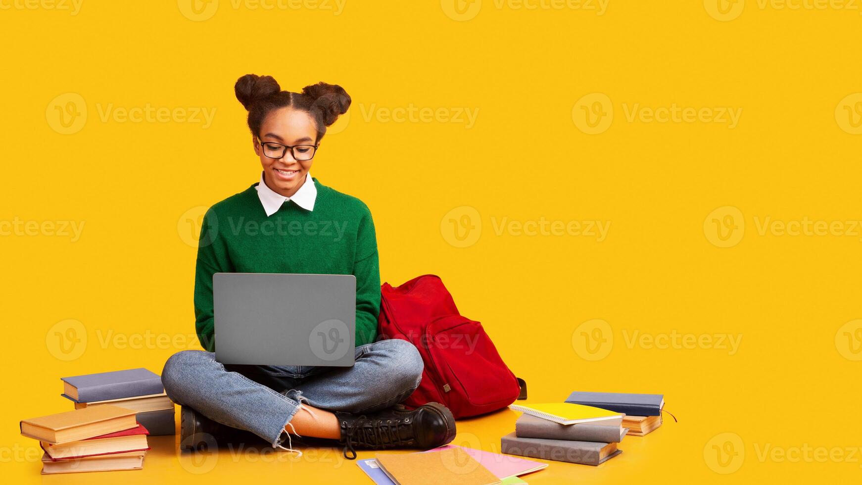Using Laptop. Happy black teenage girl doing homework, sitting on the floor, yellow studio background, copyspace photo