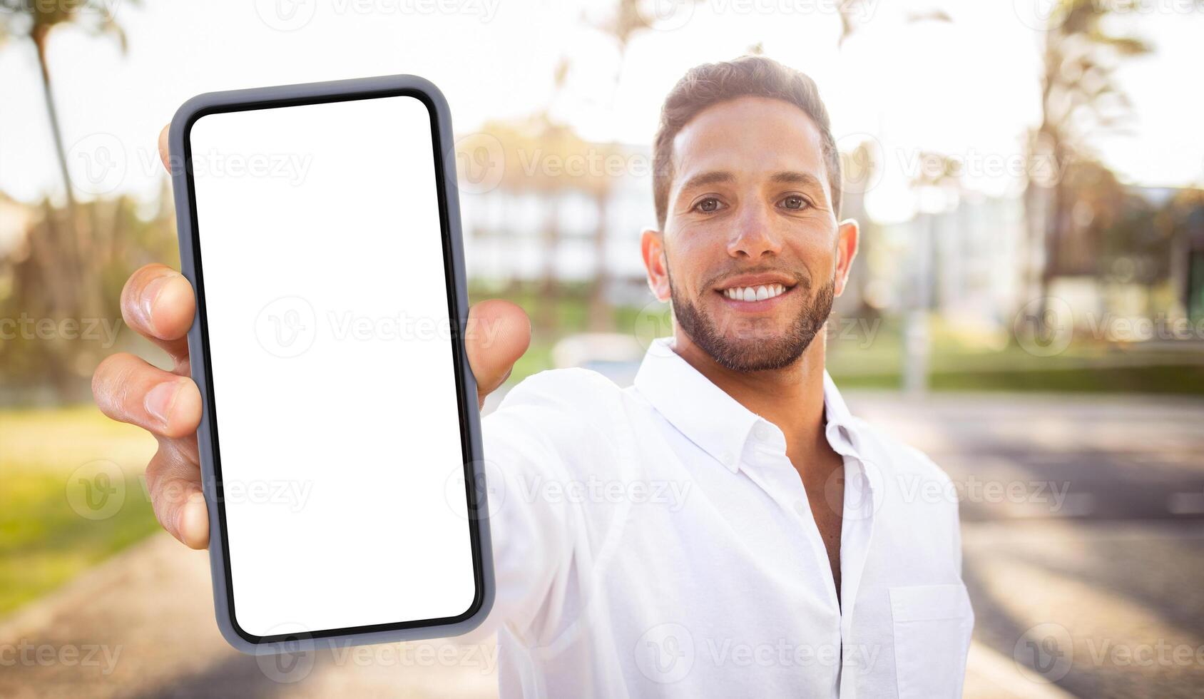 A smiling man in a white shirt holds a smartphone with a blank screen in front of him. The phone is in focus, while the man is blurred in the background. photo
