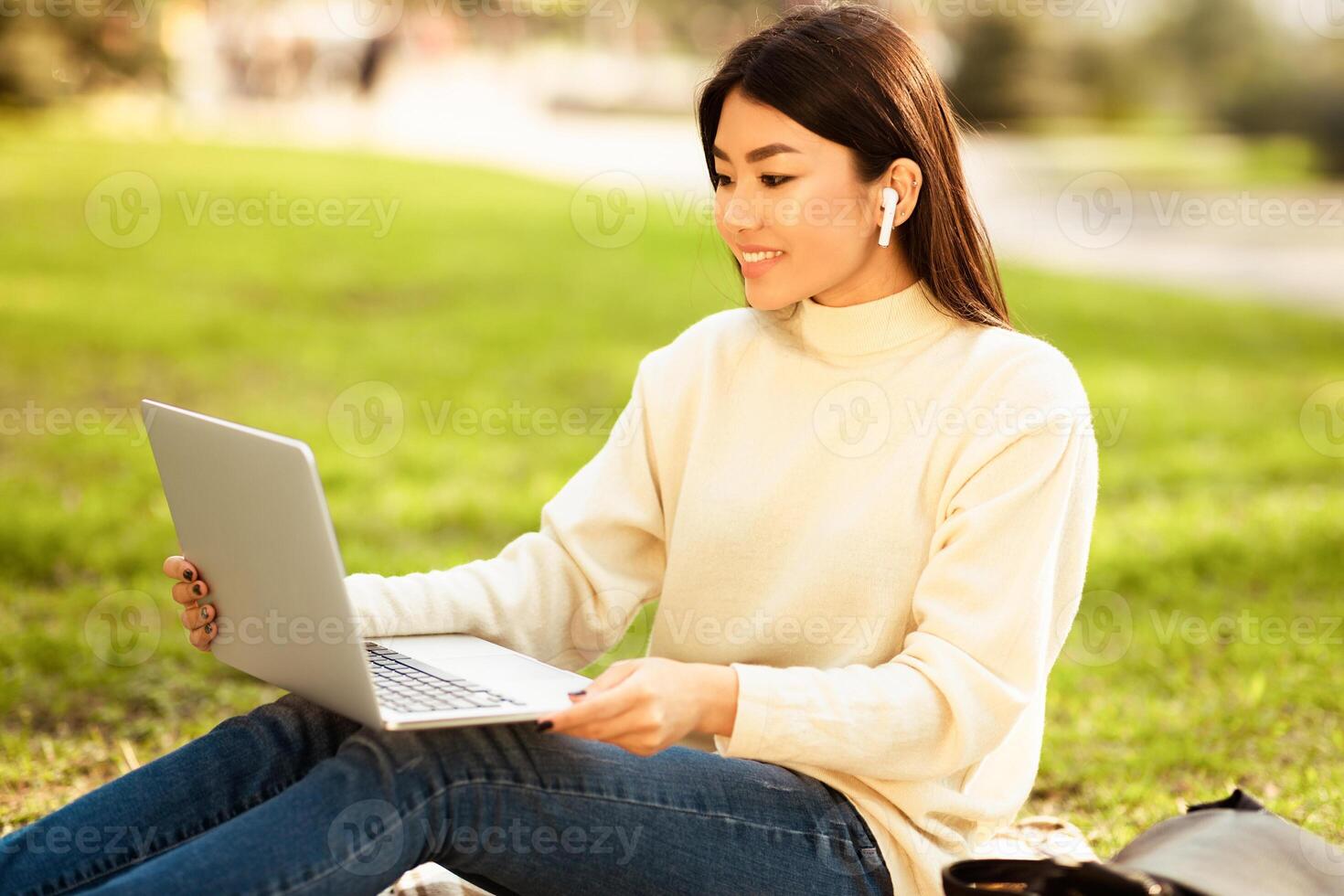 Asian woman is seated on green grass, focusing on her laptop computer and earbuds. She is typing on the keyboard, engrossed in her work. photo