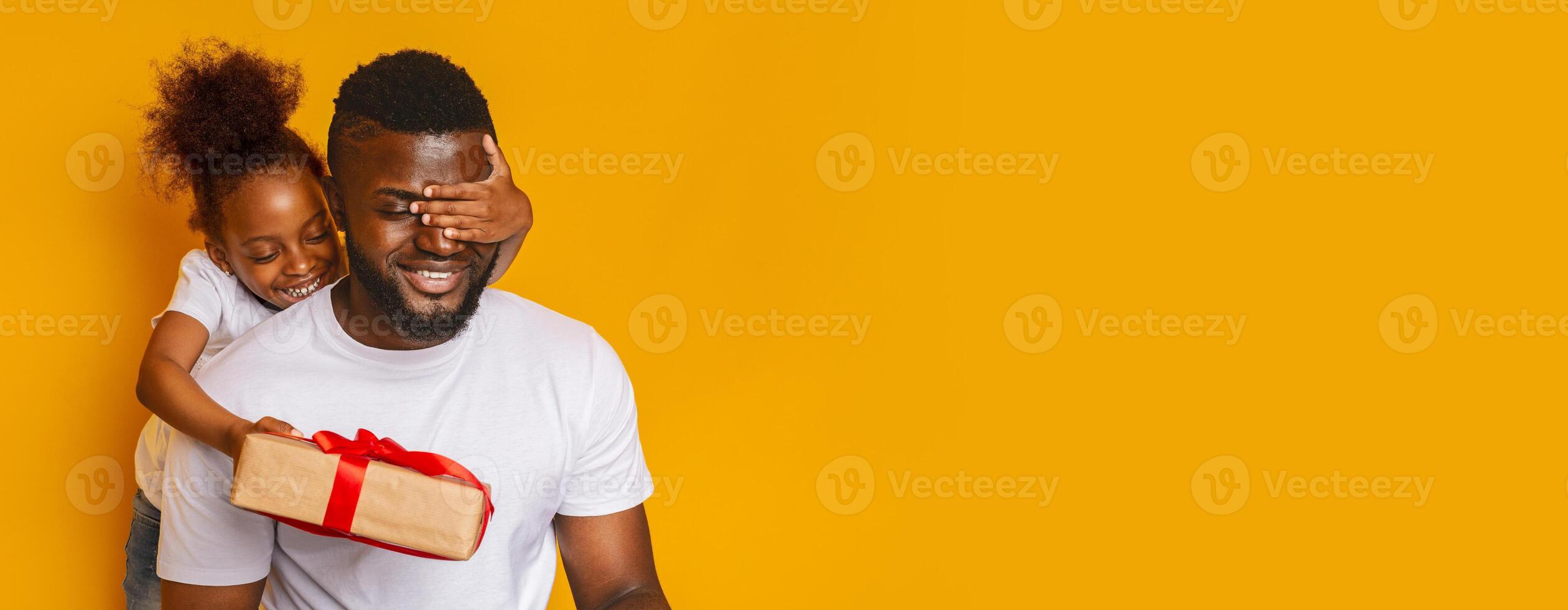 A young black girl hides her father's eyes from behind while playfully preparing to reveal a gift. The father displays a broad smile, surrounded by vibrant yellow, creating a cheerful atmosphere photo