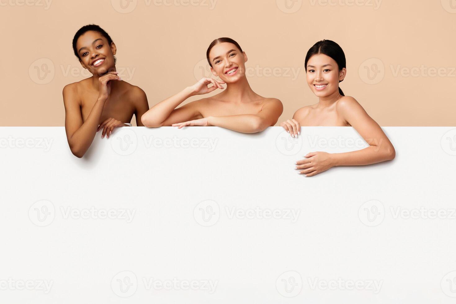 Three Multicultural Girls Posing Behind White Board With Copy Space Standing Over Gray Background. Studio Shot photo