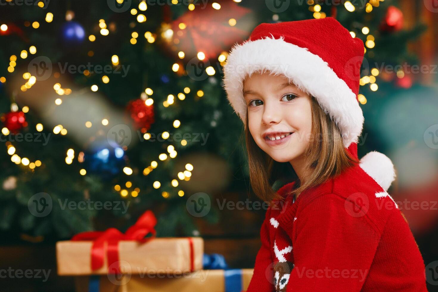 Cute preschool girl in santa hat feeding teddy bear with cookies and milk next to xmas tree photo