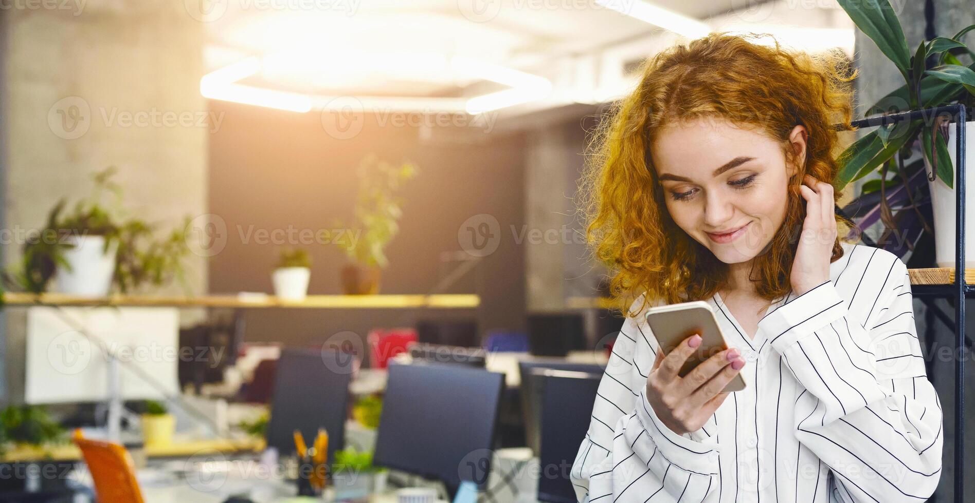 A woman is sitting in an office, focused on her phone screen, reading or typing a message. The office desk is cluttered with papers and a computer monitor is visible in the background. photo