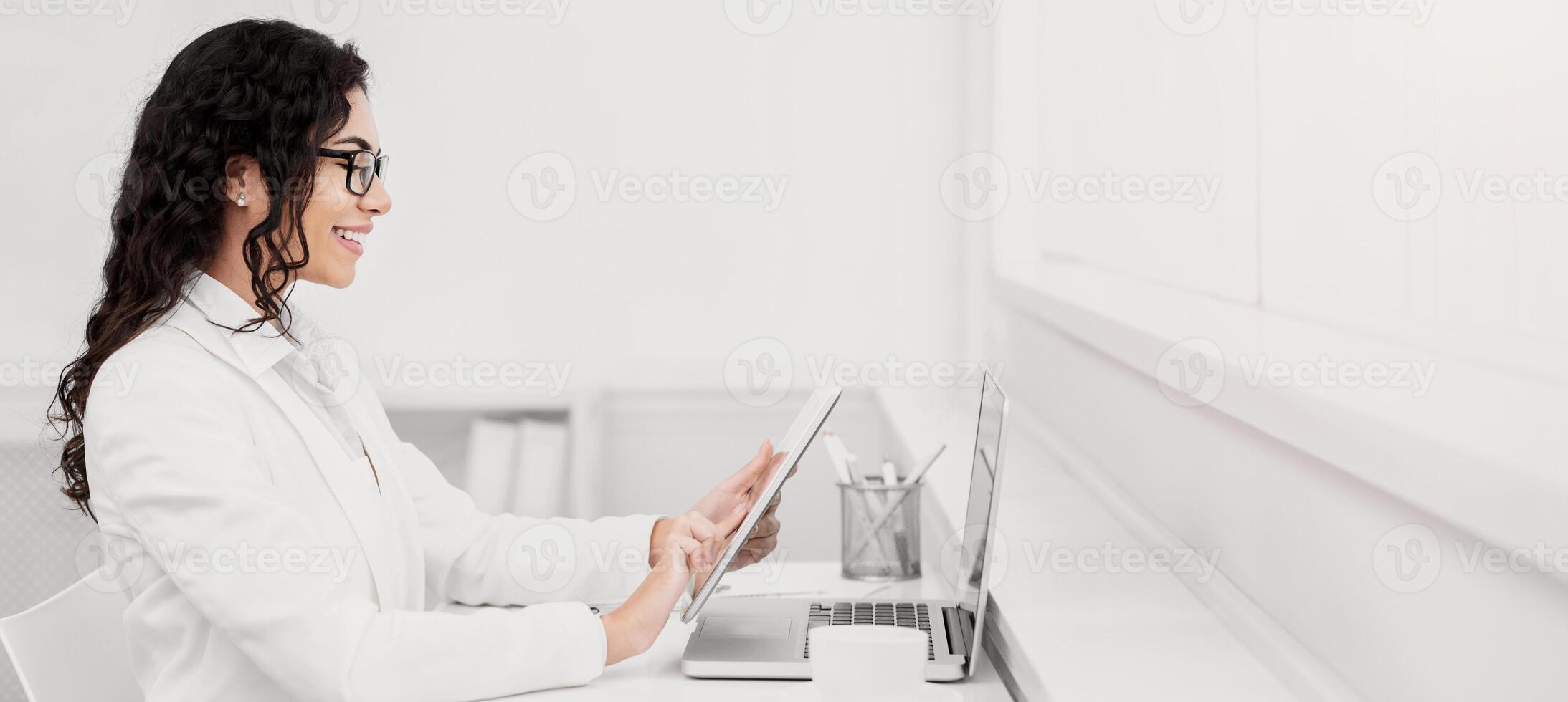 Break Time. Side view of mexican woman reading story on e-book at work, holding tablet in front of laptop, copy space photo