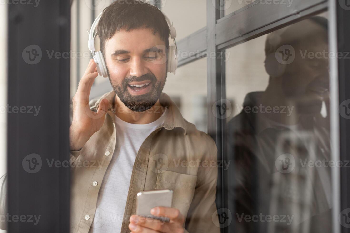 A man with a beard is singing along to music playing on his smartphone through headphones. He is standing near a window, and his reflection is visible in the glass. photo