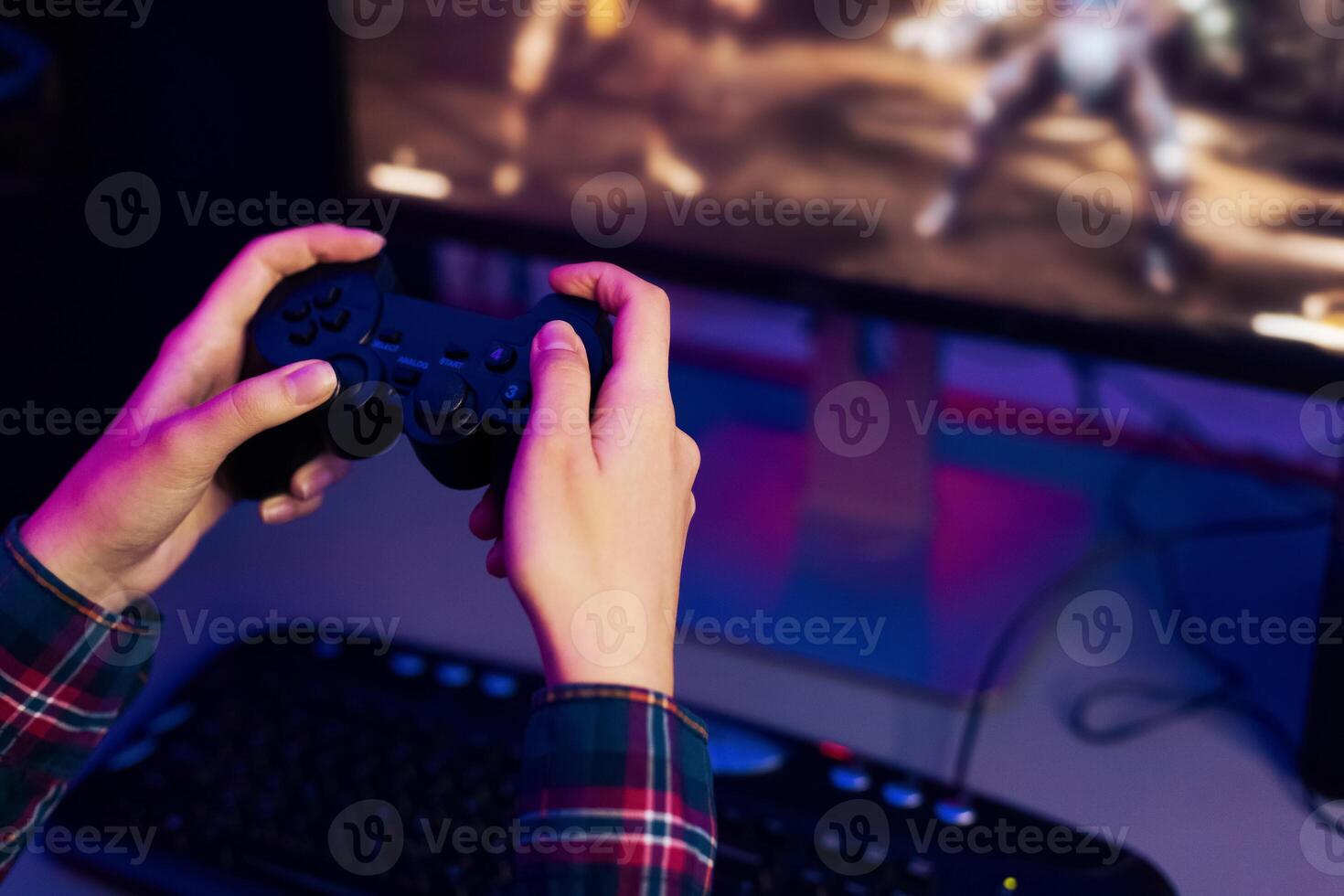 This is a photo of a persons hands holding a game controller, with a computer monitor in the background displaying an action game. The person is immersed in the game, focused on the screen.