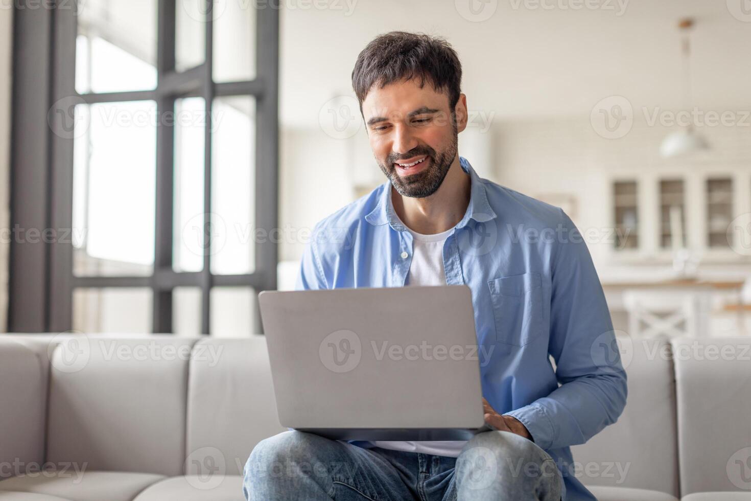 A man sits on couch in a modern home, smiling as he uses a laptop computer. He is dressed casually in a blue shirt and jeans. The home is bright and airy, with large windows letting in natural light. photo