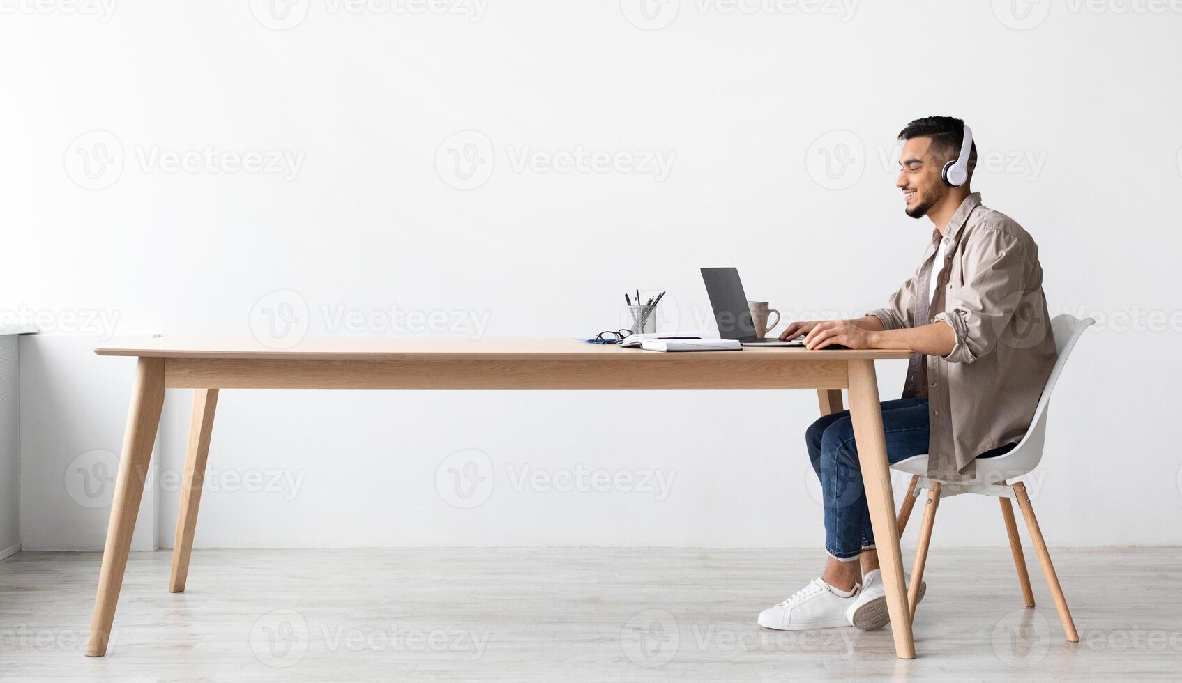 Cheerful Middle Eastern Freelancer Man Working On Pc Online At Home Listening To Music With Wireless Headphones, Making Research For New Project, Sitting At Desk Enjoying Remote Job, Profile Side View photo