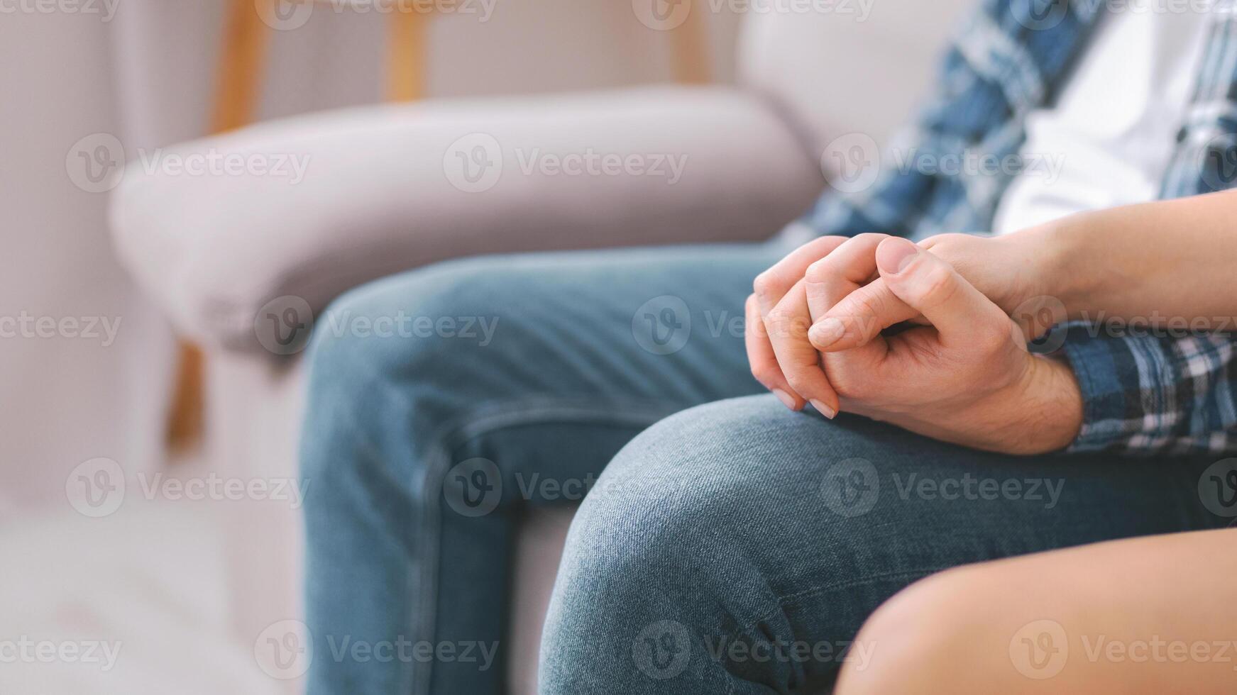 A close-up shot of a couples hands clasped together while they sit on a couch indoors. The image focuses on the intimacy of the moment and the connection between the two individuals. photo