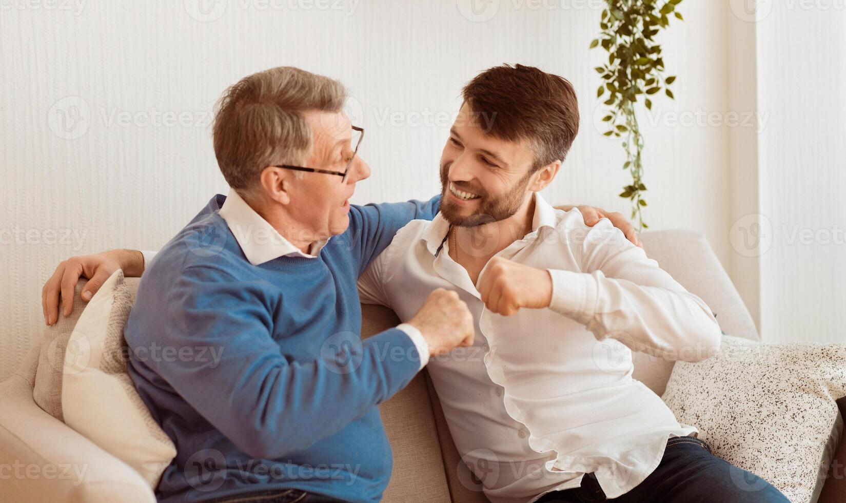 Adult Son And Elderly Father Bumping Fists Sitting On Couch At Home. Relationship With Senior Parents. Selective Focus photo