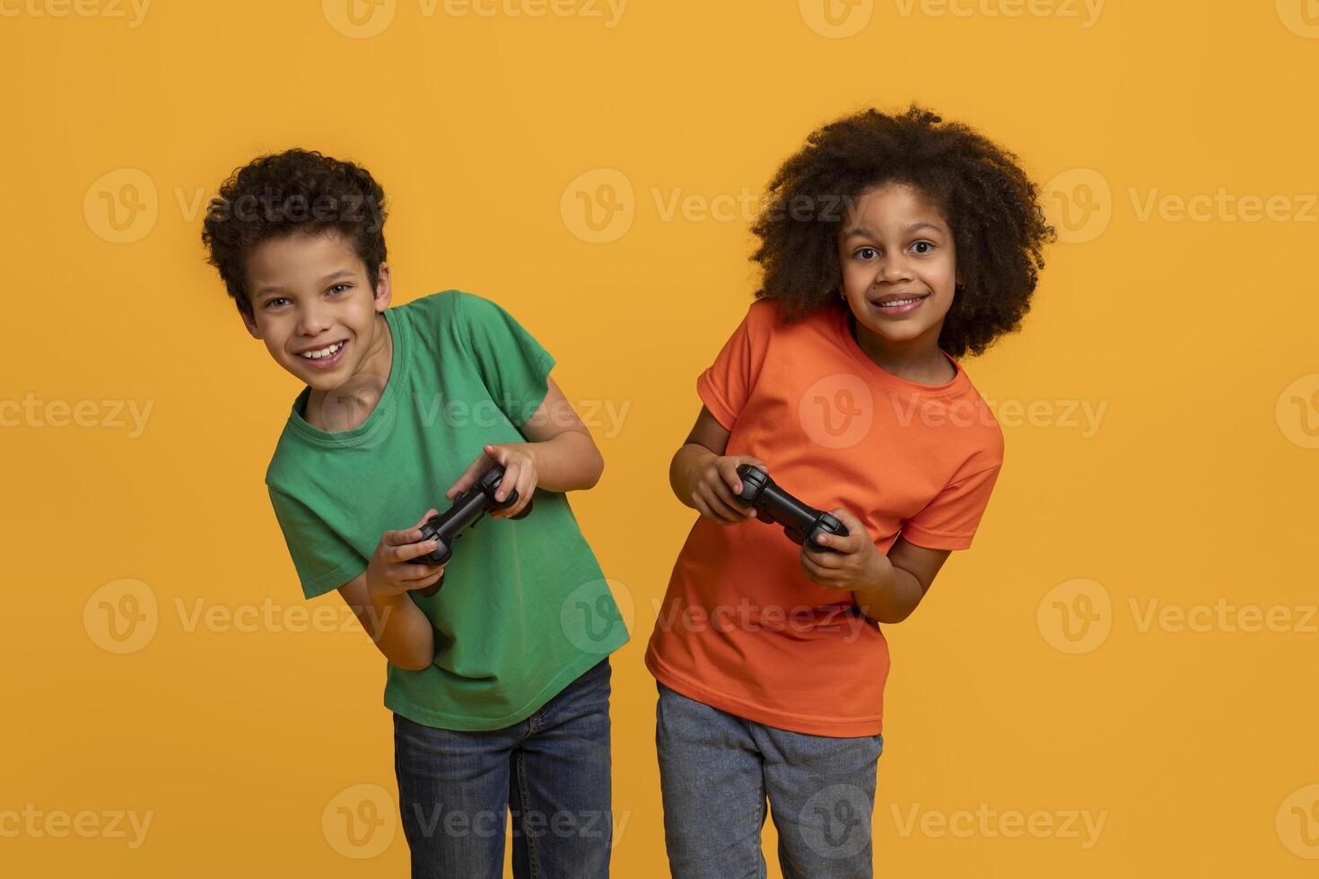 Two African American young children, a boy and a girl, stand side by side with big smiles, each holding a game controller, yellow background photo