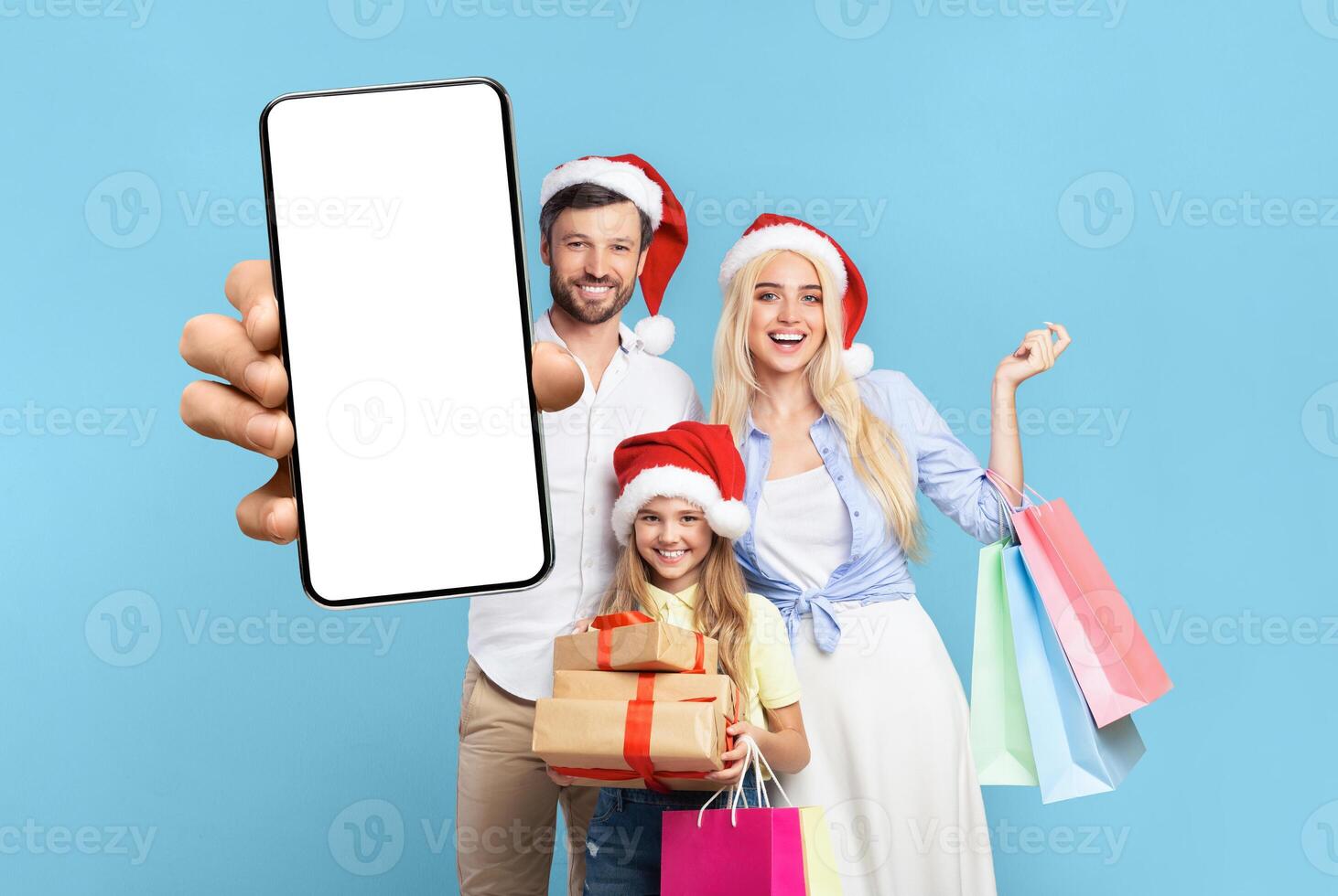 A family of three, a man, a woman, and a young girl, are all wearing Santa hats. They are standing in front of a blue background. The man is holding a smartphone in front of him photo