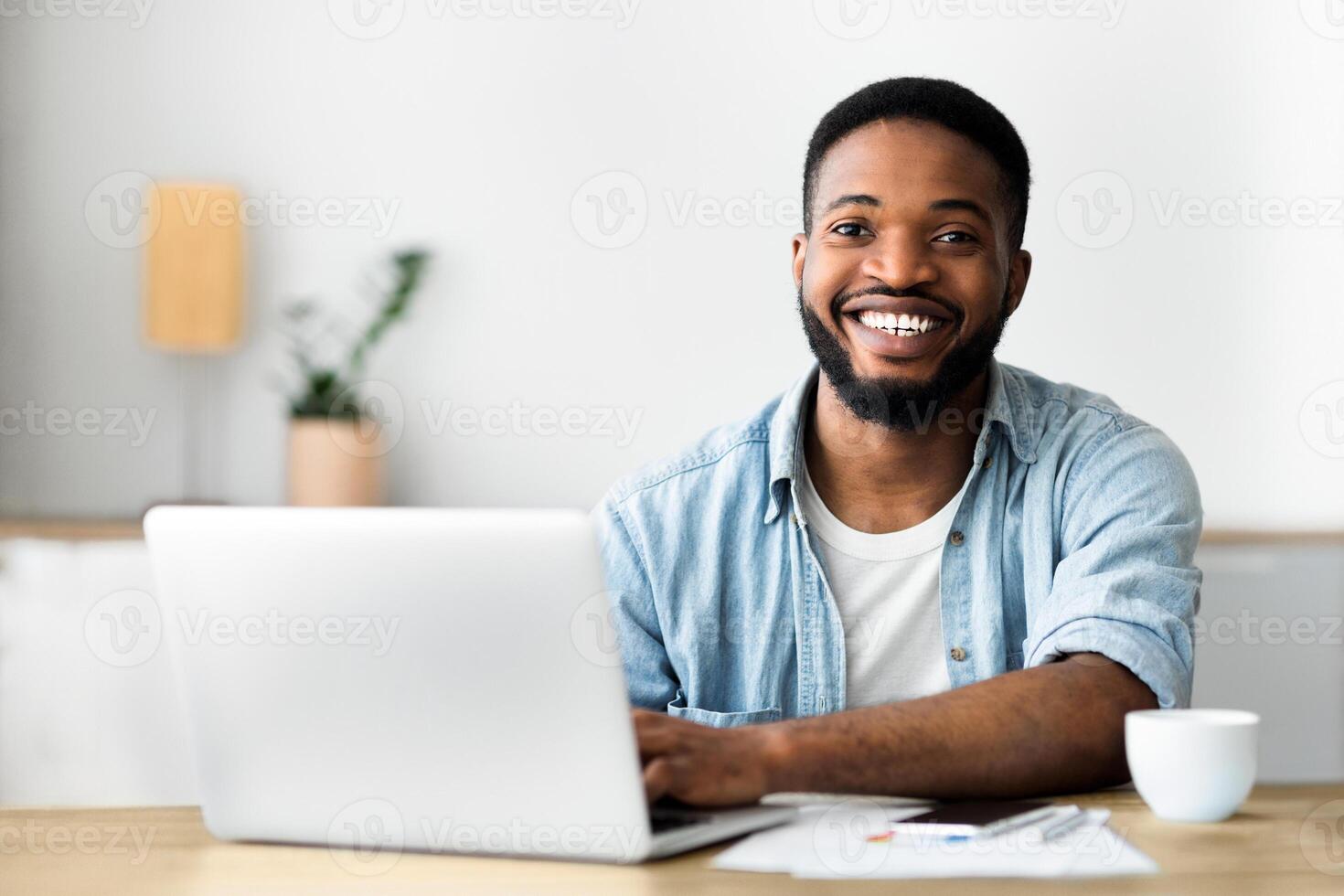 Portrait of handsome african worker using laptop, smiling and drinking coffee at workplace in modern office. Free space photo