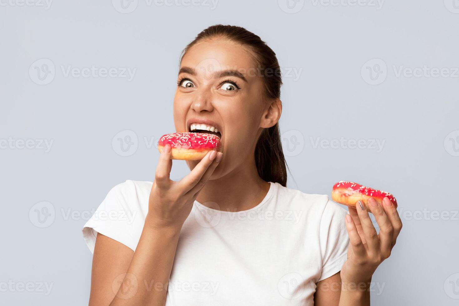 Hungry Girl Eating Donuts Standing Over White Studio Background. Unhealthy Sweets Concept photo