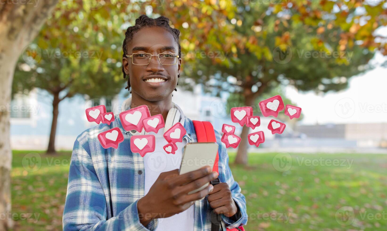 A young black man wearing a plaid shirt and glasses stands in a park with a backpack, smiling as he checks phone. Digital hearts and notifications float around him, suggesting social media activity. photo