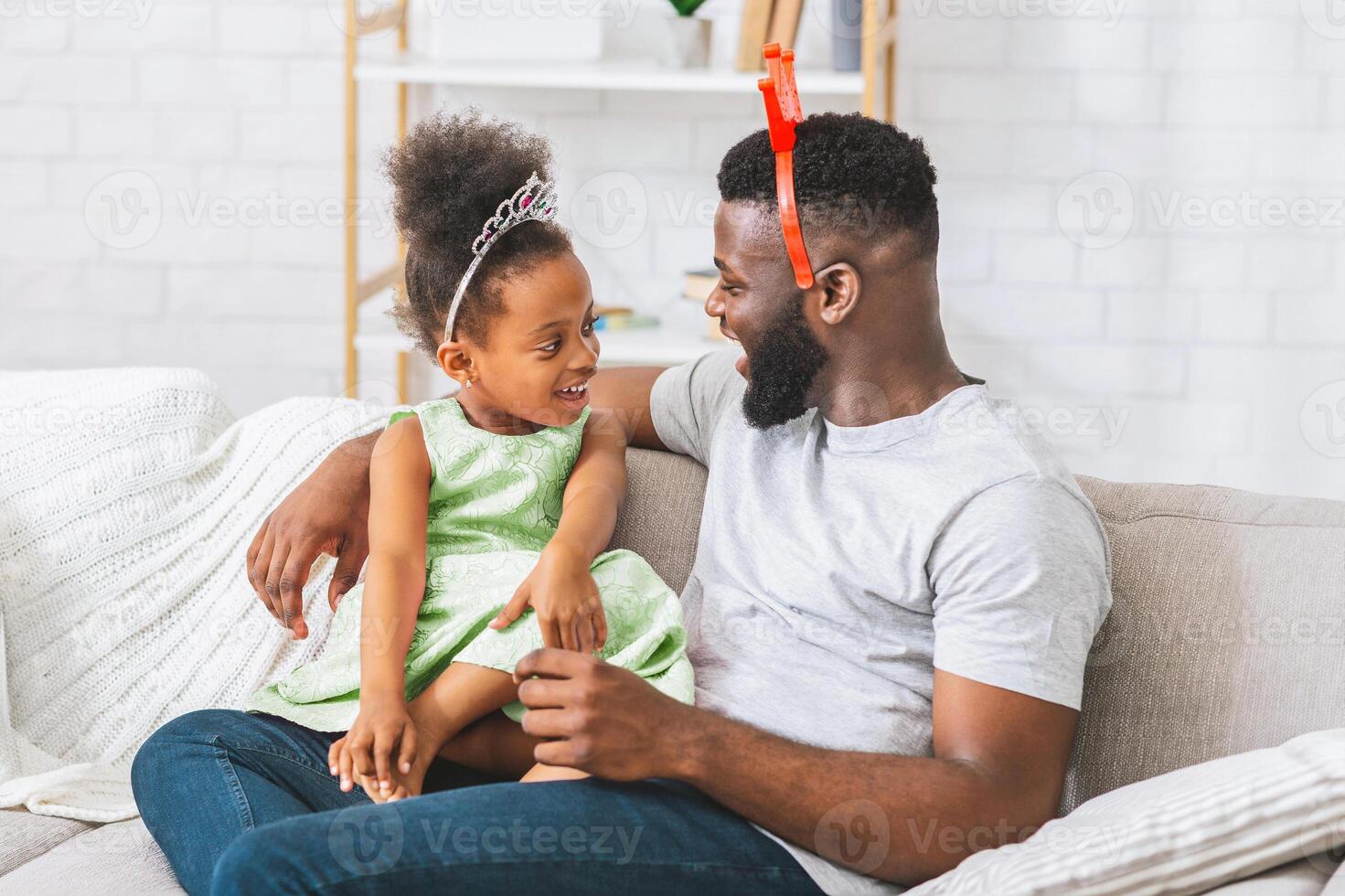 A father and daughter are sitting on a couch together. The daughter is wearing a princess crown and a green dress. The father is wearing a white T-shirt. Both are looking at each other and smiling. photo