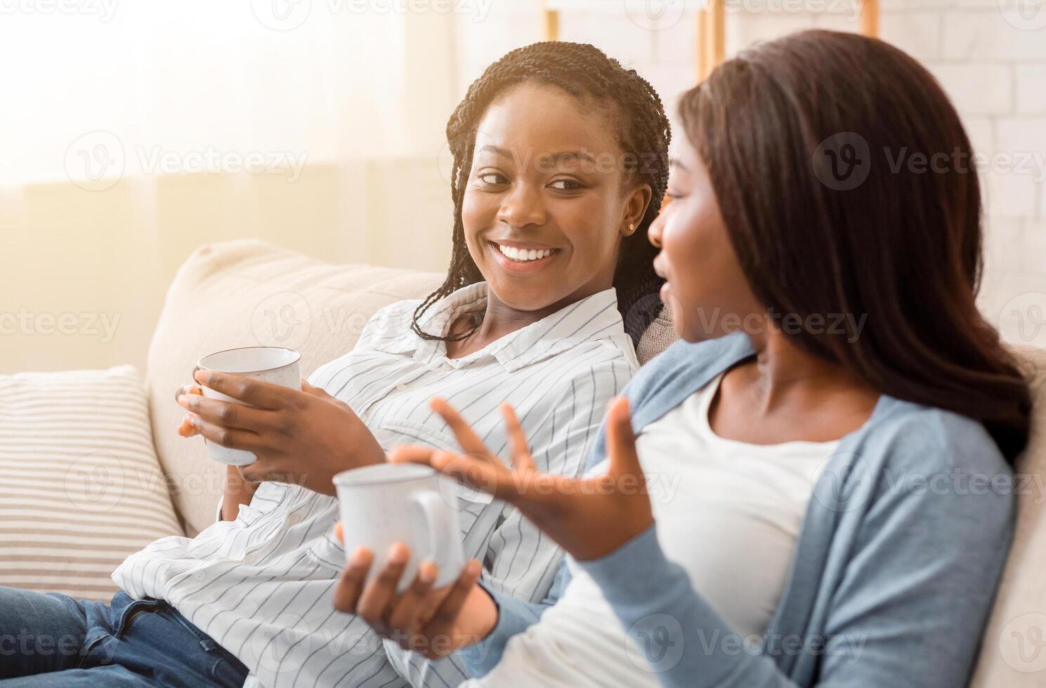 Two female friends sitting together on sofa chatting and drinking tea, having fun at home. photo