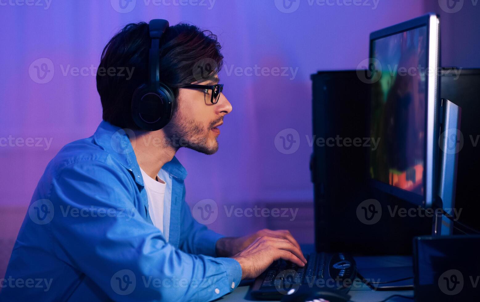 A man is sitting in front of a computer with headphones on, playing a game. He is focused on the screen and his hands are on the keyboard. The room is lit with purple lighting photo
