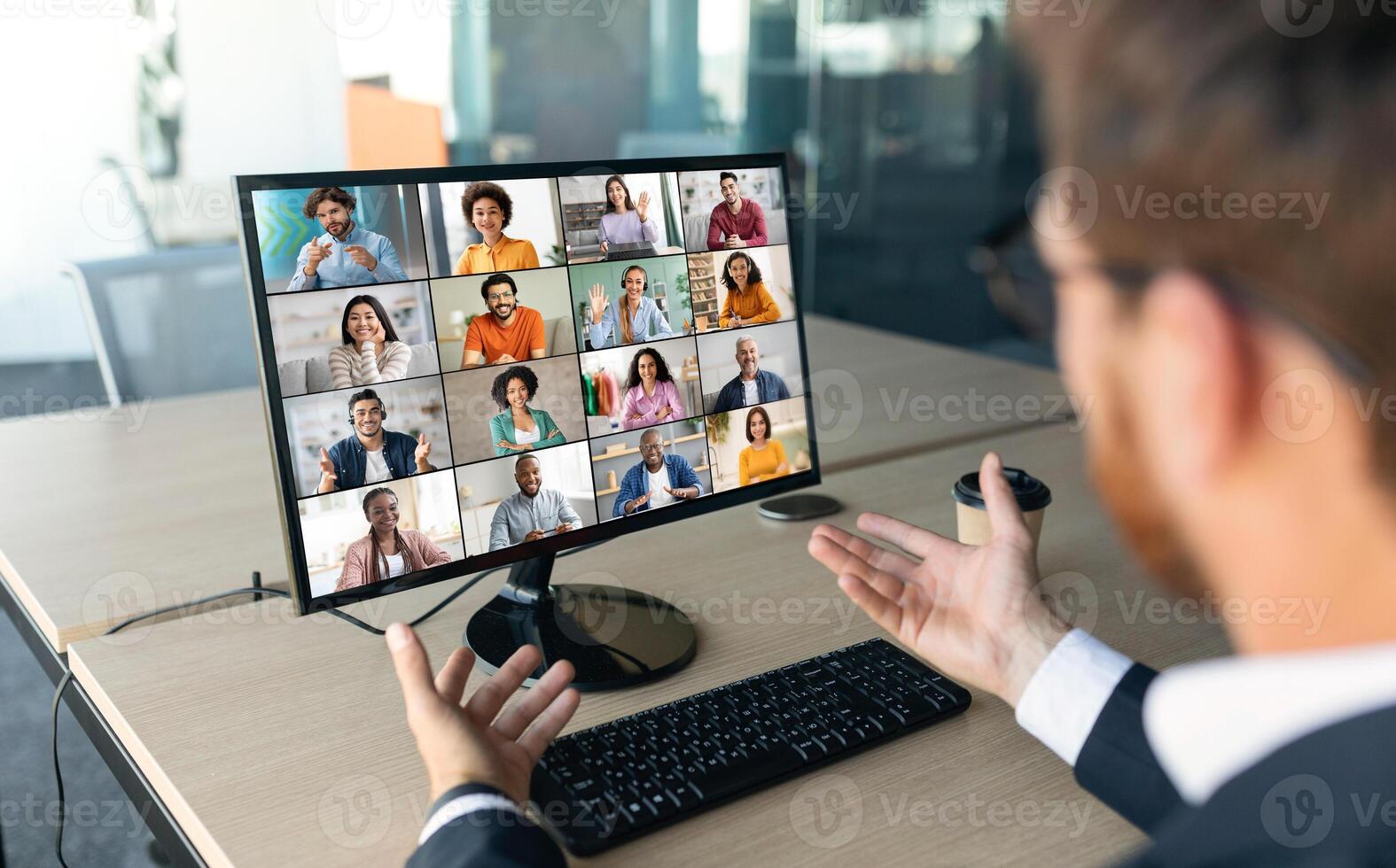 A businessman sits at a desk in his office, with a computer screen displaying a conference call with several other people. He is gesturing with his right hand photo
