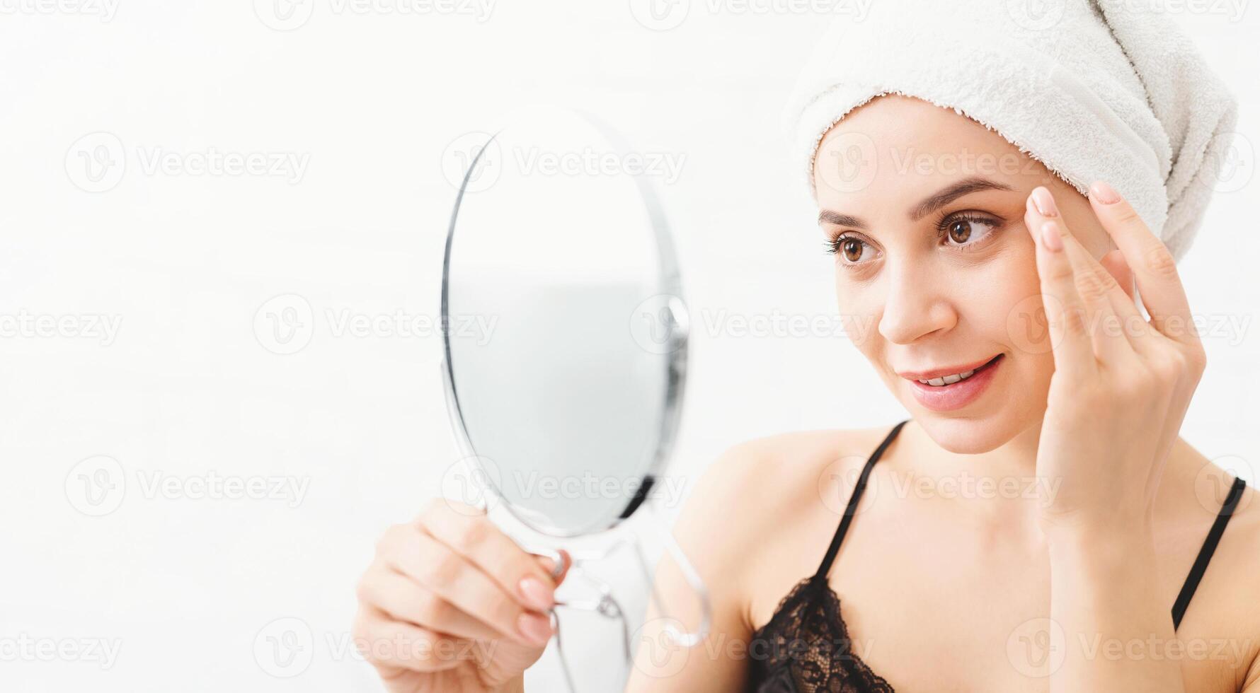 A woman with a towel wrapped around her head is focused on examining her face in a mirror. She is likely checking her skin condition or applying skincare products. photo