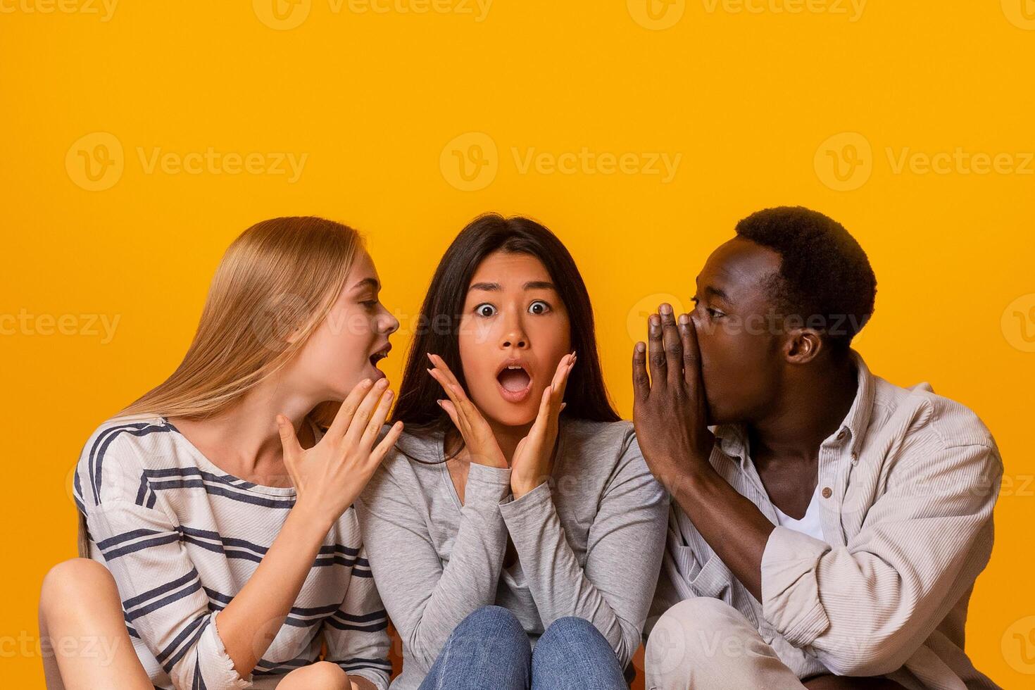 International friends sitting in row on floor, gossiping over yellow studio background, student lifestyle photo
