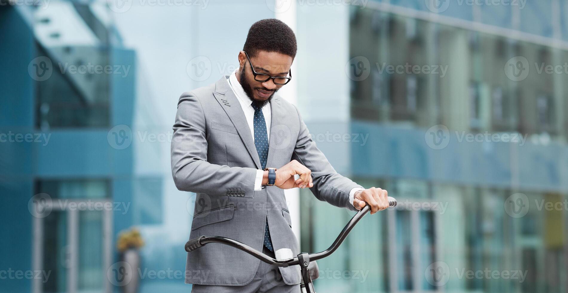African American businessman on a bike in the city is checking his watch. The setting is urban and busy, capturing the essence of multitasking and staying on schedule in a fast-paced environment. photo